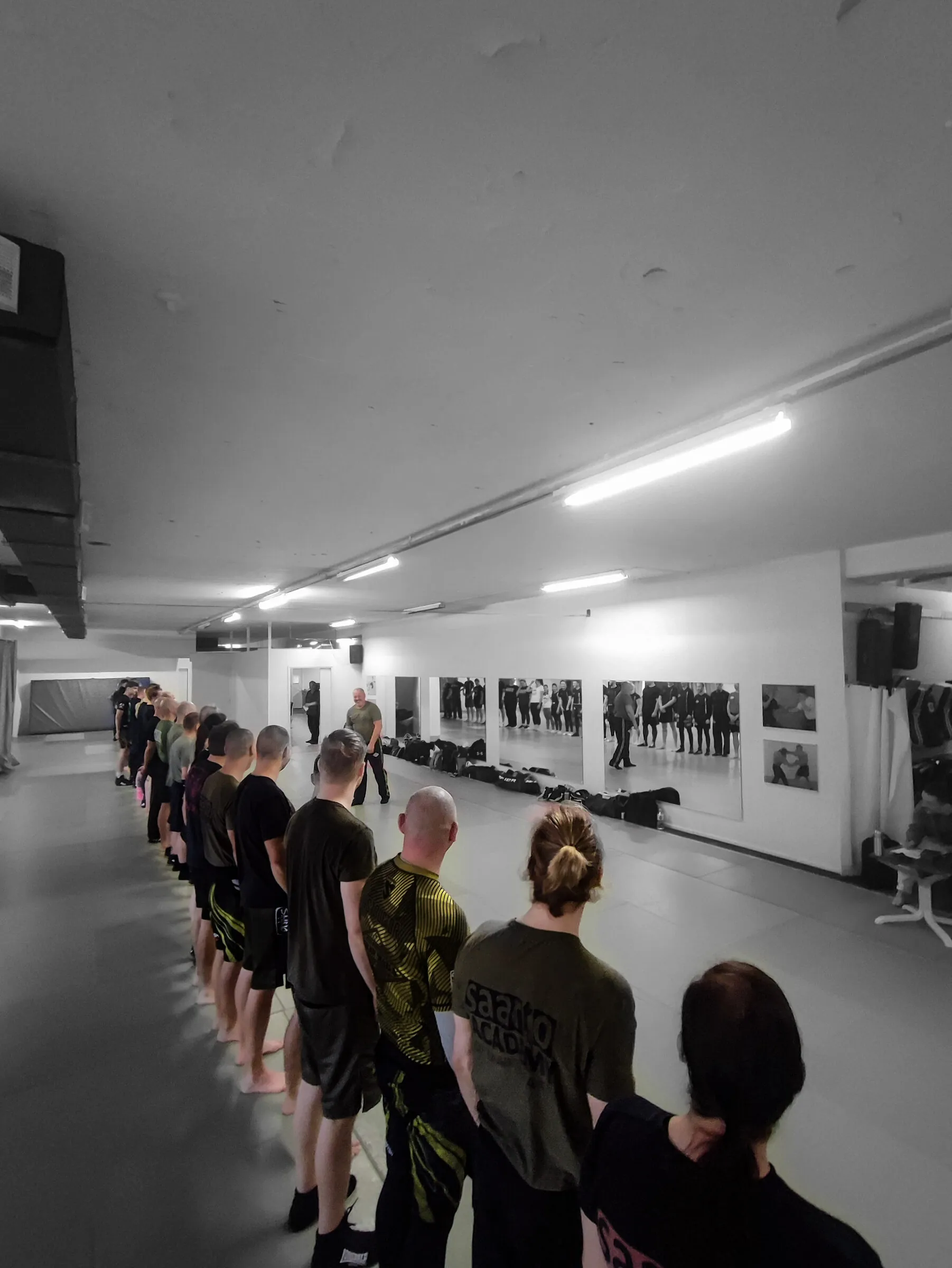 A course line of practitioners standing at attention on the mats, backs to camera, as the lead instructor addresses them across the training hall
