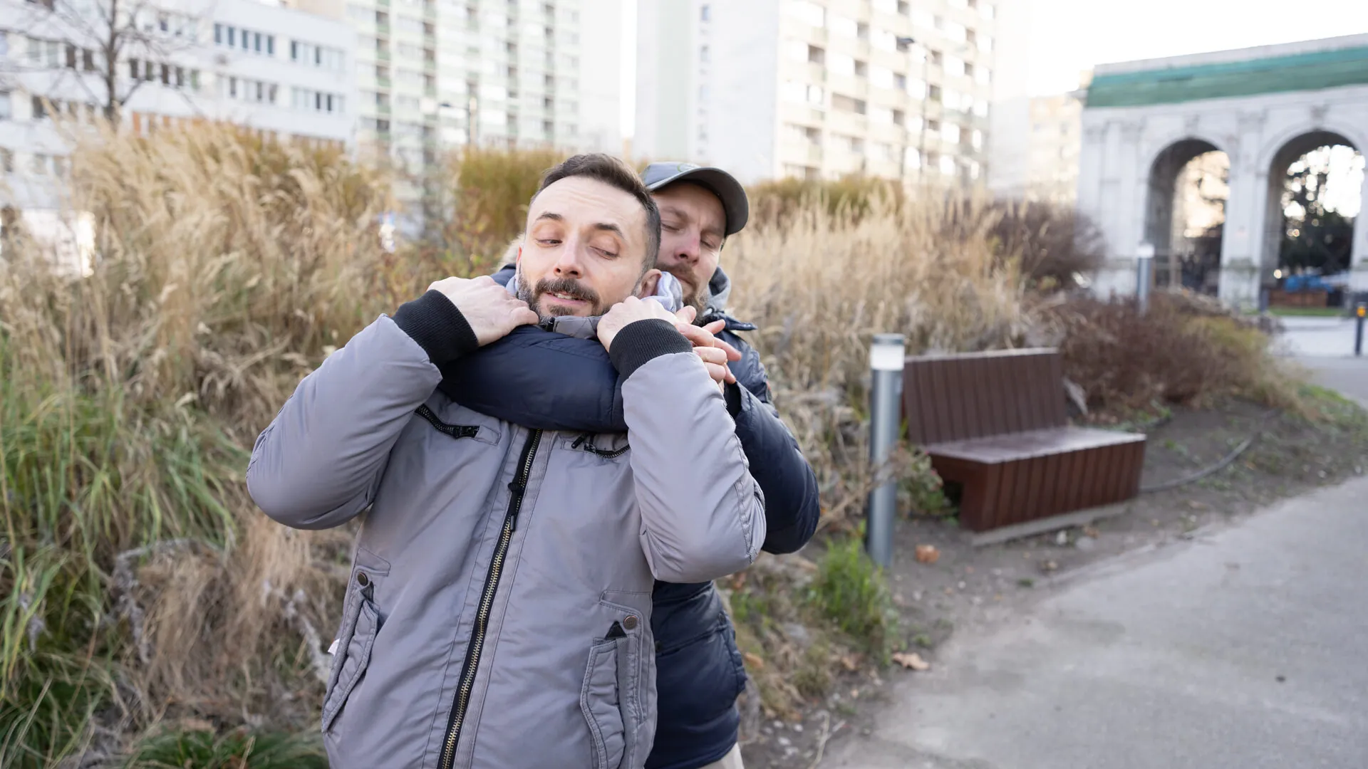 A man defends against a rear choke on a Warsaw street, dry winter grasses and apartment blocks behind