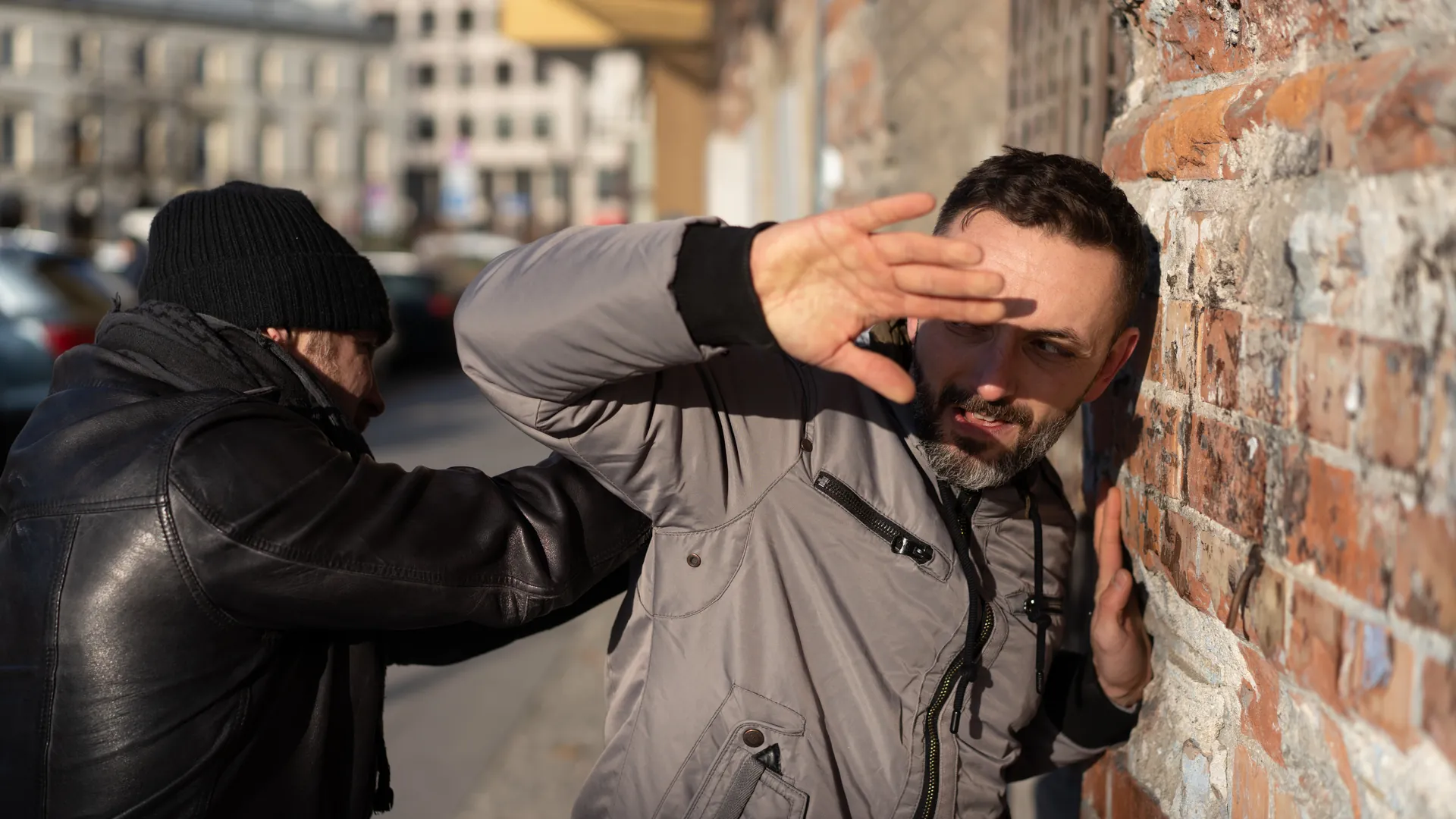 Street-situations training in Warsaw — a man raises an open hand to shield his head against a sudden attack at a brick wall, city street behind