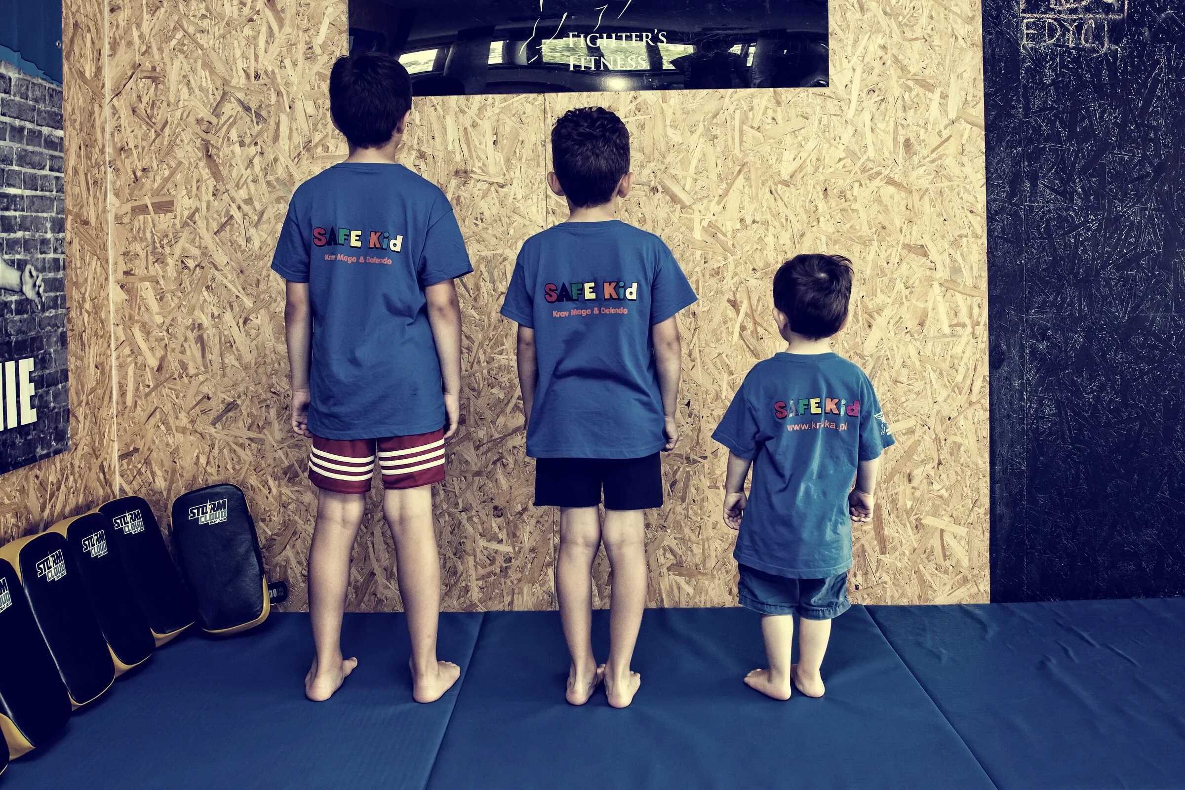 Three children in blue Safe Kid shirts standing on the mats, seen fully from behind, facing the gym wall
