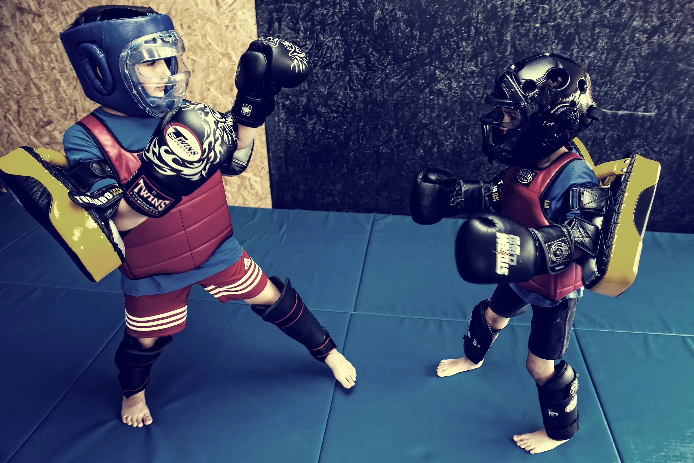 Two young students spar in full protective armour — helmets, gloves and pads — on soft mats