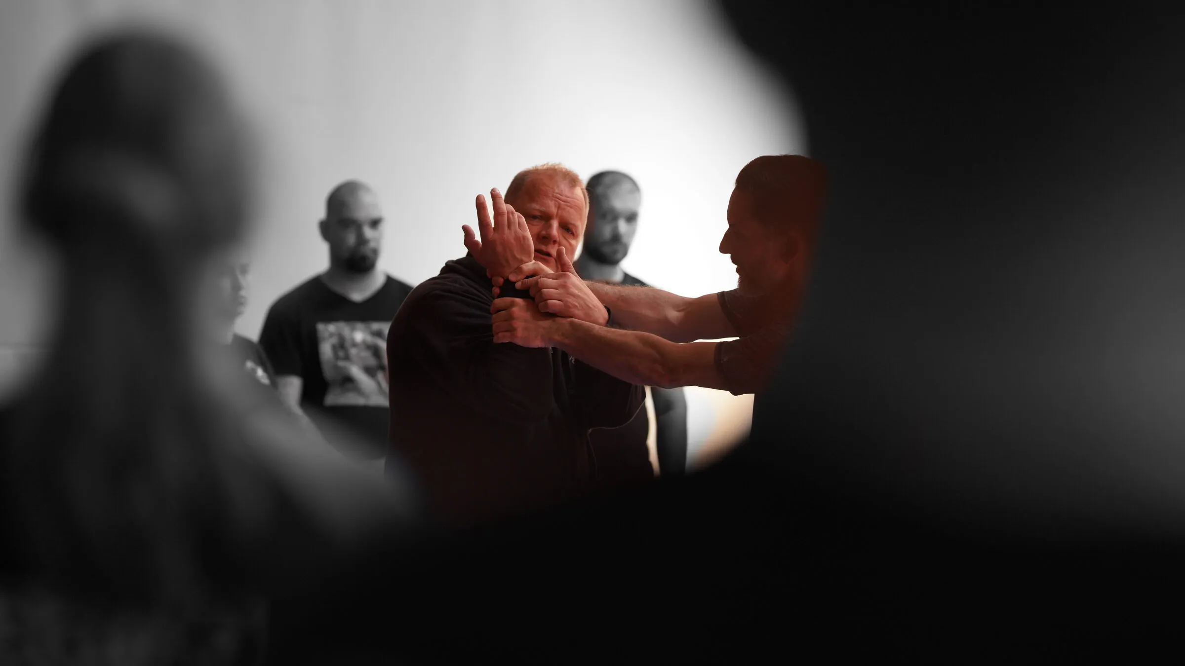 A seminar seen through blurred foreground heads — a senior instructor demonstrating a control while the class watches