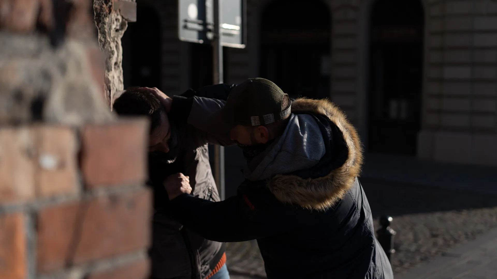 A deep-shadow street scenario against a brick wall — one man pinning another, the urban reality the professional programs train for